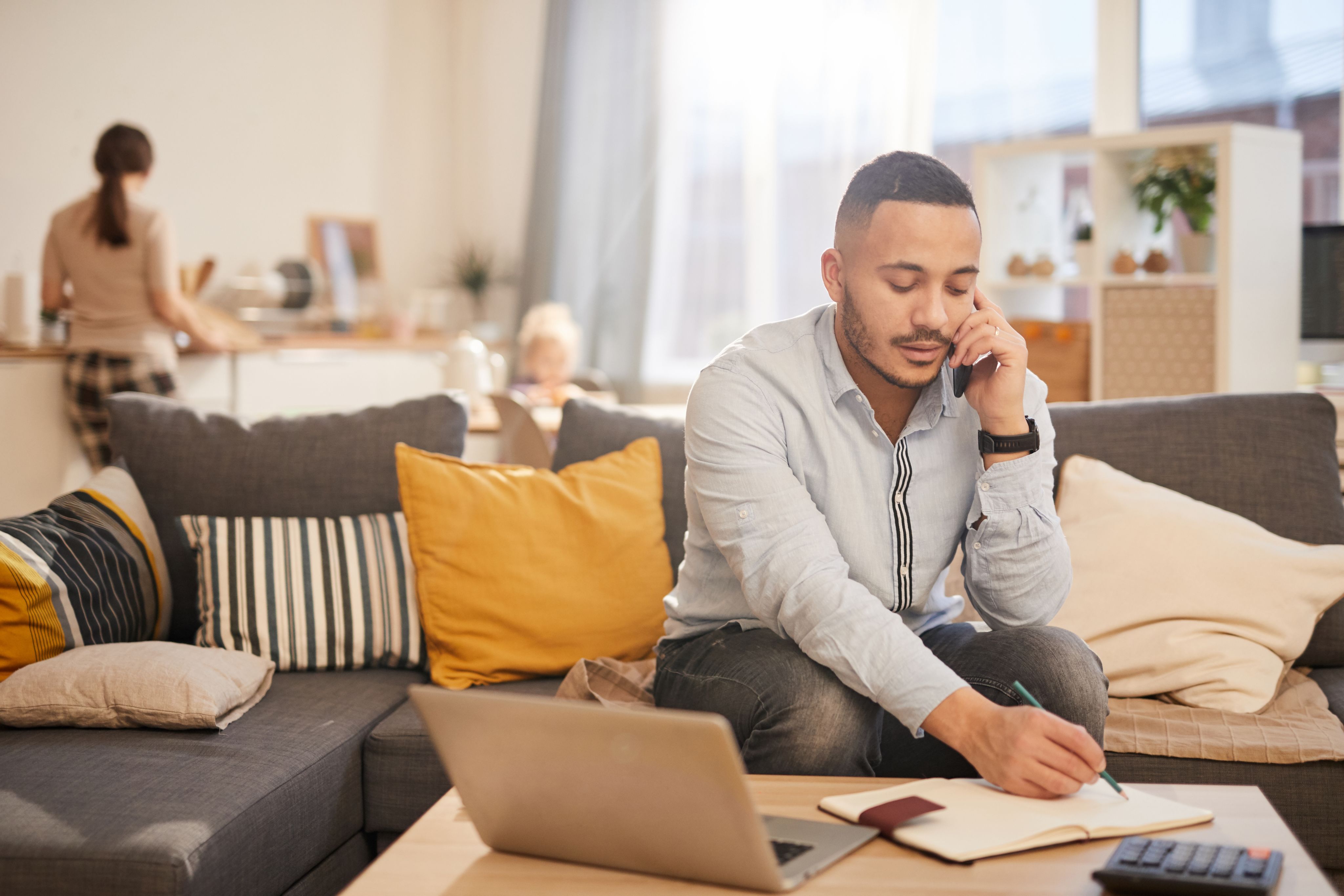 A man works from home on the phone on his couch while his partner is in the kitchen in the background.